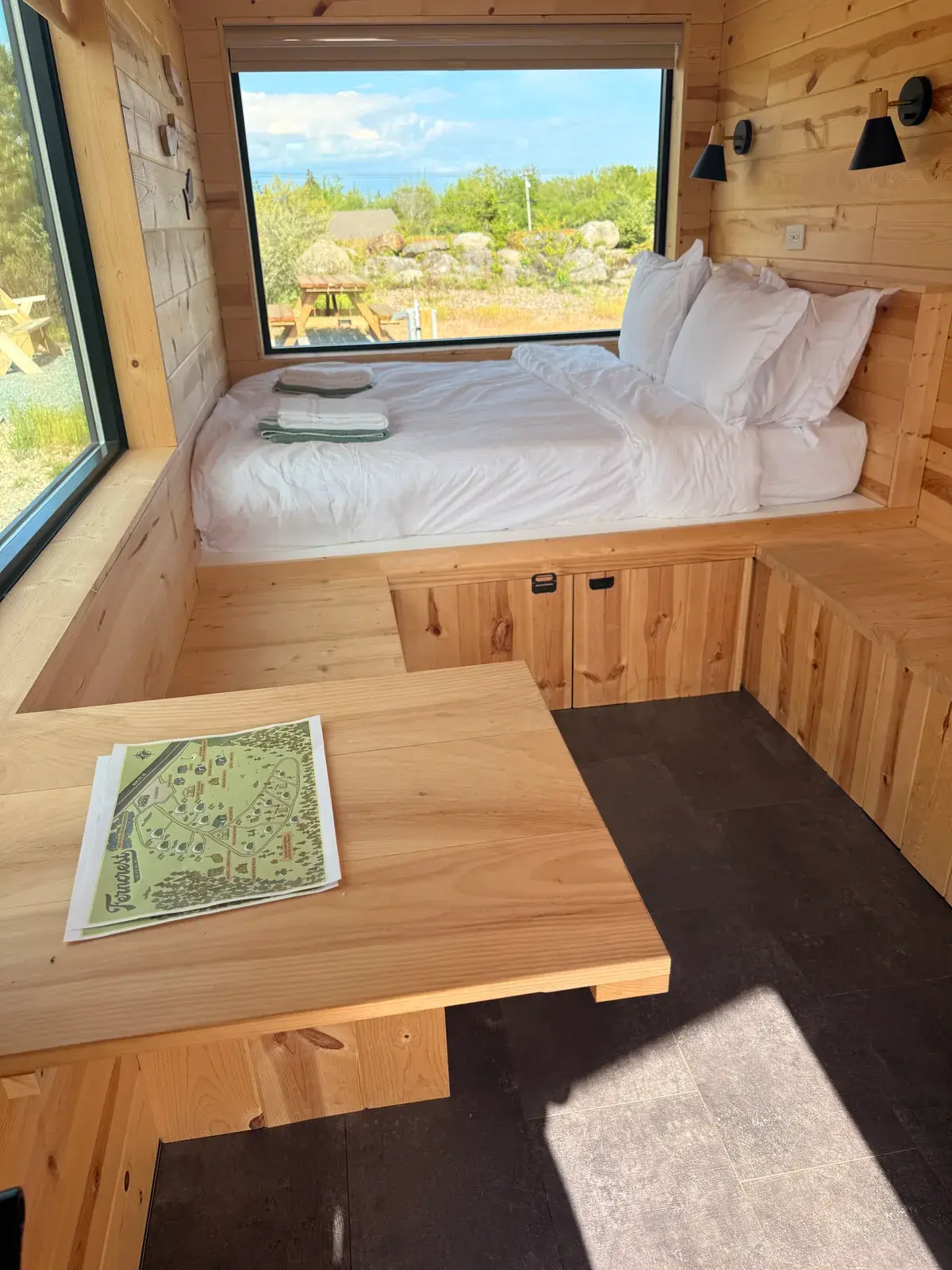 Cabin interior with a made bed beside a large forest-view window